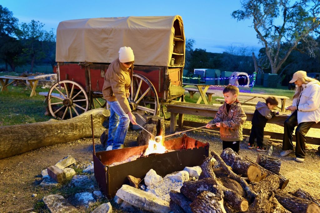 A family roasting s mores at a campfire beside the covered wagon at Old West Christmas Light Fest